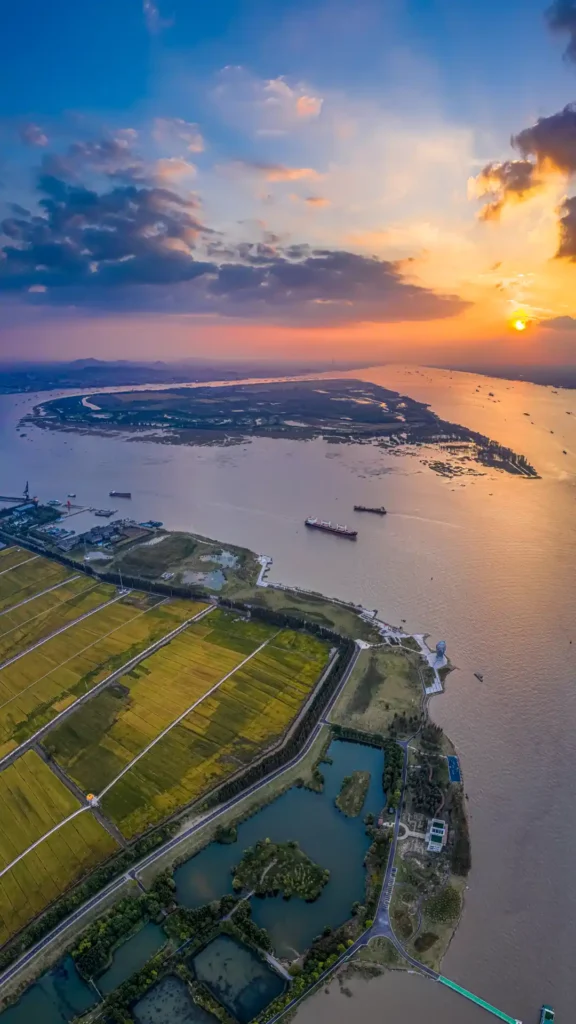 Aerial vertical panoramic view of a riverside landscape at sunset: golden agricultural fields, scattered ponds, and winding roads line the wide river, where cargo ships navigate; a river island stretches in the distance, under a sky with orange-blue gradient clouds and a glowing setting sun.