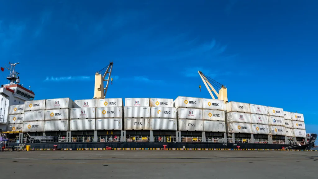 A container cargo ship docked at a port, loaded with stacked white shipping containers marked with "IRNC", "ITSS" and other logos, equipped with yellow cranes on board, set against a bright blue sky with wispy clouds, showcasing a busy international trade logistics scene.