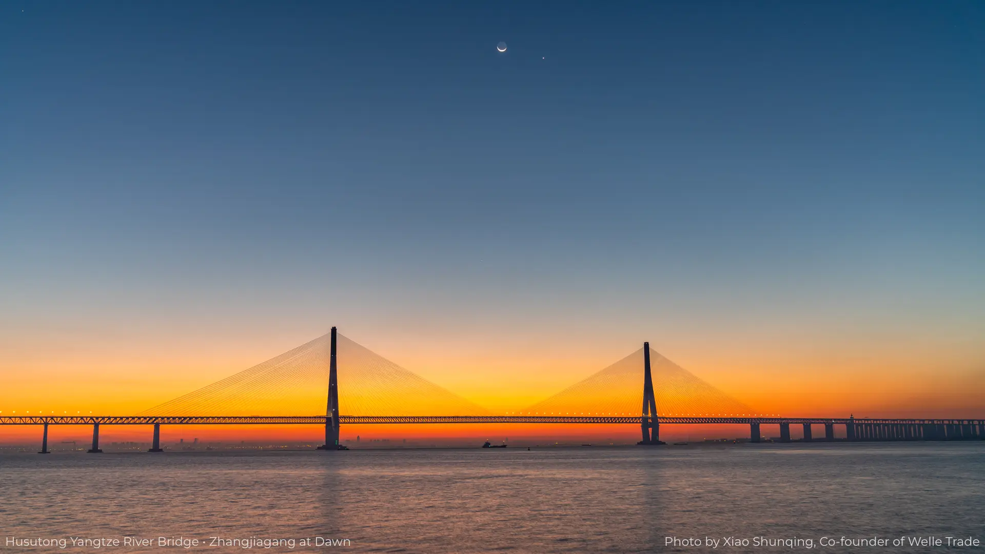 Dawn view of the Husutong Yangtze River Bridge in Zhangjiagang: a cable-stayed bridge spanning calm water, silhouetted against a gradient sky of orange and blue, with a crescent moon and a star visible above, and small ships on the river; this photo is taken by Xiao Shunqing, co-founder of Welle Trade.