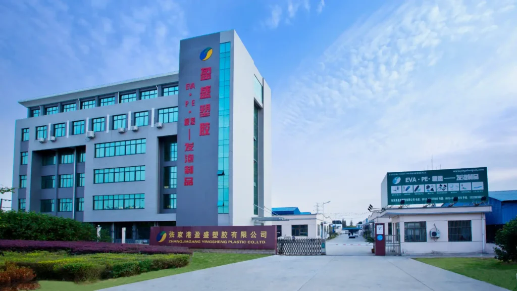 Exterior view of Zhangjiagang Yingsheng Plastic Co., Ltd. office building: a modern multi-story structure with gray-white facade and glass windows, featuring vertical red Chinese characters "Yingsheng Plastic" and "EVA,PE Foam" on the side, a company name sign at the entrance, a product billboard on the right, set against a blue sky with white clouds and neat greenery around.